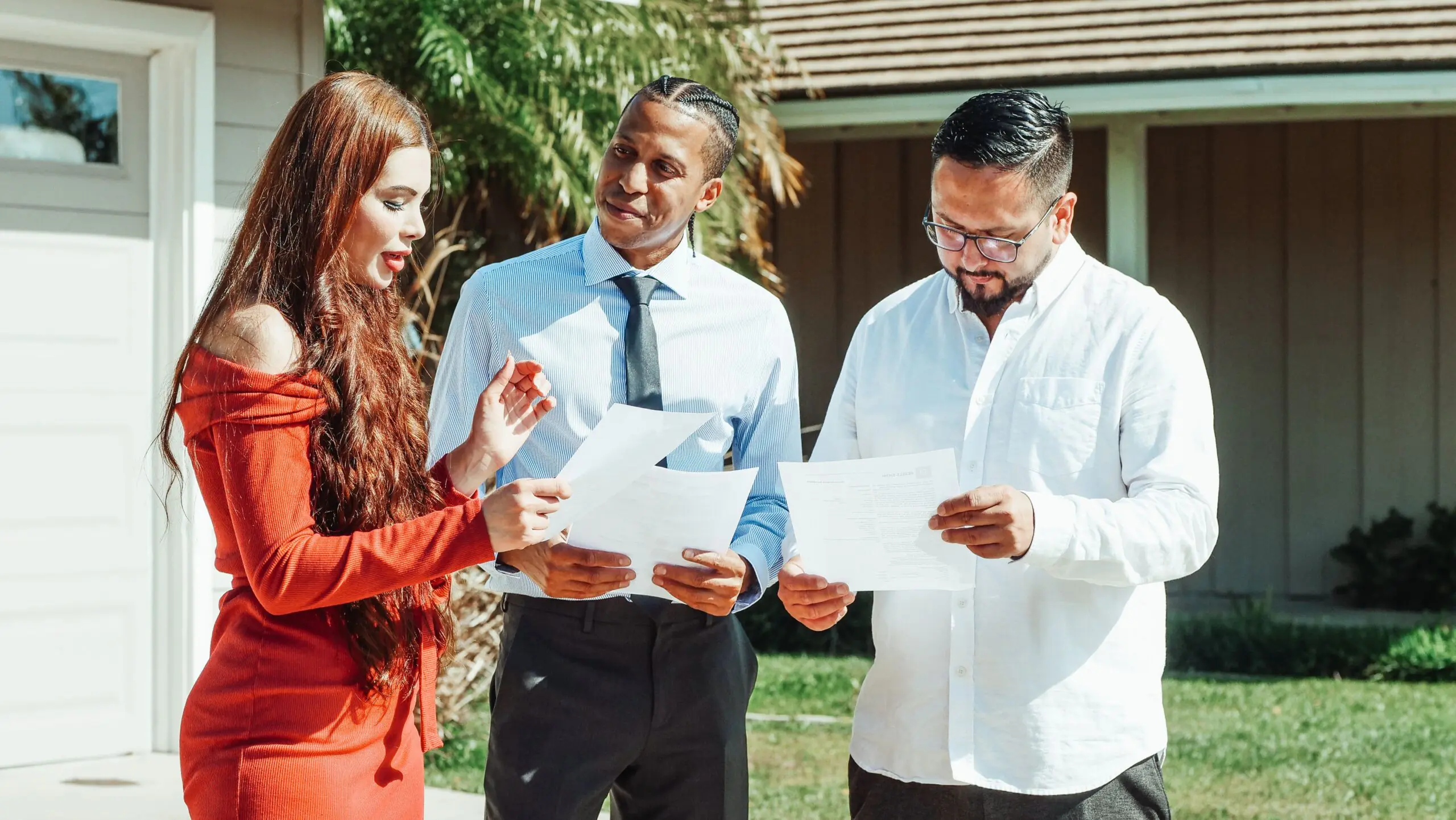 A diverse group evaluates real estate documents outside a residential property in daylight.