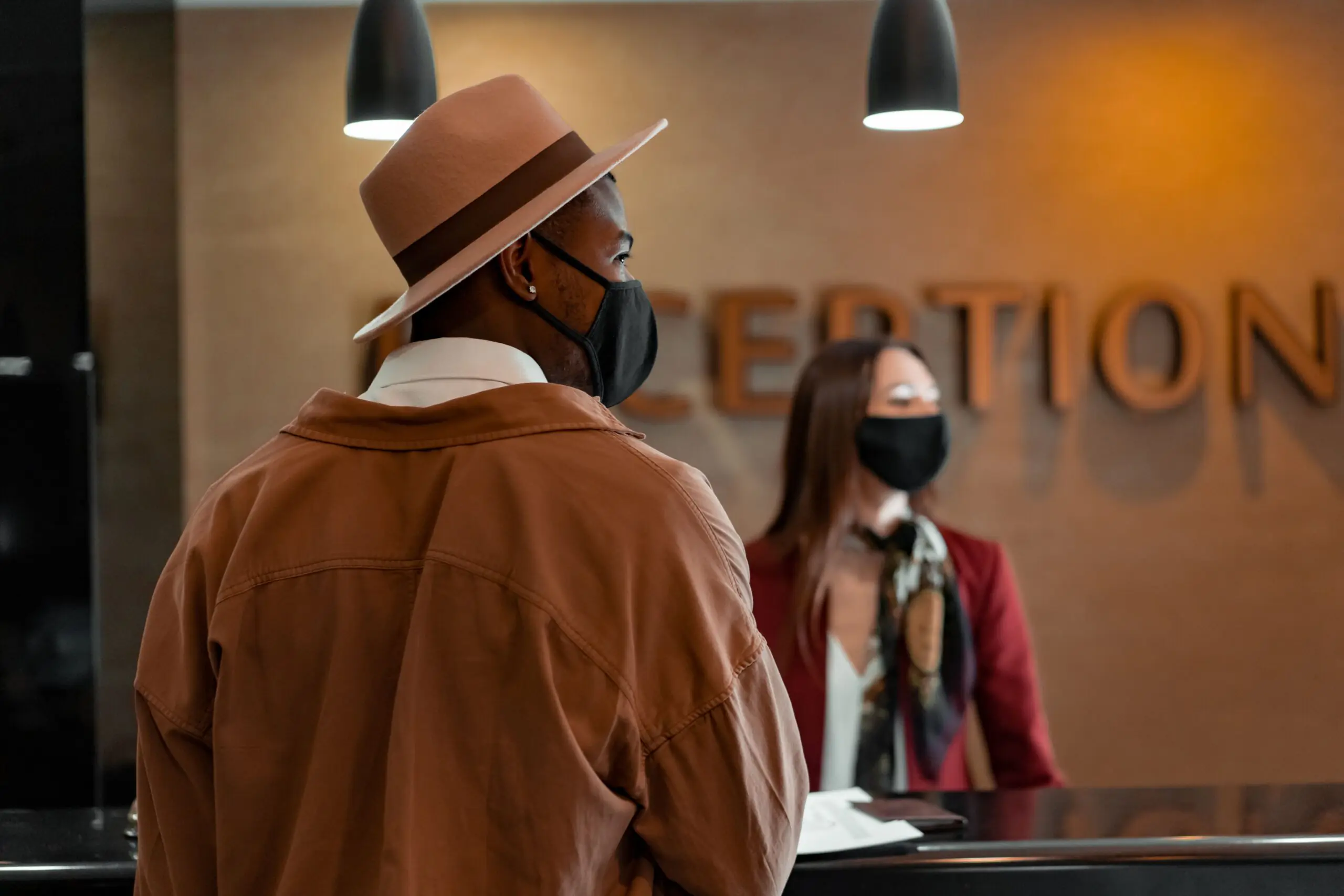 Man in mask at hotel reception during pandemic, interacting with masked receptionist.